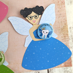 A paper cut fairy in a blue dress holding a 'I lost my first tooth' badge