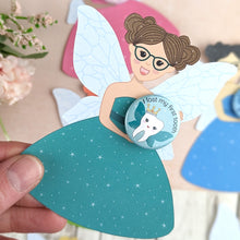 Load image into Gallery viewer, A hand cut tooth fairy with brown hair and dark jade glasses. She is wearing a jade dress with sparkles and has pale blue wings. She is holding a I lost my first tooth badge..