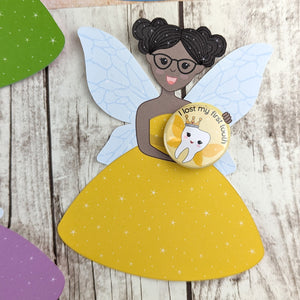 A tooth fairy wearing a yellow dress and black glasses. She is holding a yellow 'I lost my first tooth' badge which features a tooth with wings.