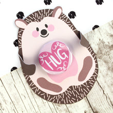 Load image into Gallery viewer, Cute pink and brown hedgehog with folded arms and a pink &#39;HUG&#39; badge